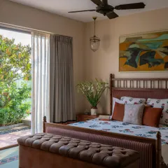 A bedroom in Calcutta opens directly onto a lush garden, blurring the line between indoors and out. The room is a calm oasis, with a beautiful wooden bed, a tufted leather bench, and a large abstract painting that adds a touch of modern energy.