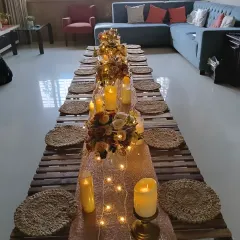 An intimate dinner party setup at home. I used a low wooden table, jute placemats, and a runner decorated with candles and flowers, all lit by warm fairy lights.