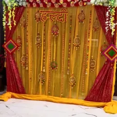 This Haldi backdrop uses a combination of sheer yellow and rich red drapes for a royal look. The decor is completed with floral arrangements at the top, a "Haldi" sign, and decorative hangings for a festive feel.