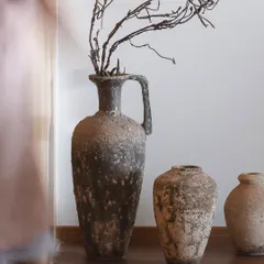 A trio of ancient-looking pottery, including a large amphora with a handle, is styled with dried branches. These pieces carry the weight of time in their texture and gentle imperfections.