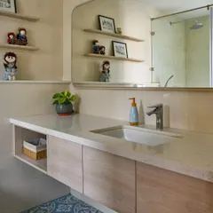 This bathroom from 'House of Kismish' features soft blue patterned floor tiles and a floating vanity with open shelves for displaying personal items. The large, custom-shaped mirror makes the space feel larger.