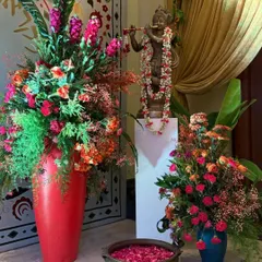 A detail of a traditional entrance, featuring a Krishna statue adorned with a floral garland. The space is decorated with vibrant floral arrangements in red and orange, and a brass 'urli' bowl with floating petals, infusing the arrival with culture and charm.