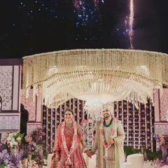 The Varmala moment, perfectly framed under a magnificent floral chandelier with fireworks lighting up the sky. This is where all the planning culminates in a single, powerful memory.