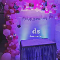 This anniversary party backdrop features a 'Happy Anniversary' sign against a simple white background, framed by a balloon arch in shades of pink and gold with butterfly accents. The purple uplighting adds a dramatic effect.
