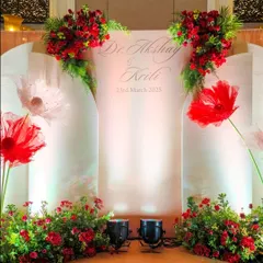 A personalized photo backdrop with the couple's name and wedding date, decorated with large, artistic paper flowers and fresh red floral arrangements.