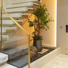 This composition combines a realistic faux tree with natural stones and polished pebbles in a contained under-stair garden. The sleek metallic planter and warm accent lighting complement the modern marble and brass staircase.