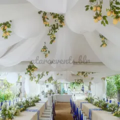 An elegant long-table setup for a Sicilian-themed event. The tables are covered in white linens with blue patterned runners, and the ceiling is draped with white fabric and hanging lemon branches, transporting guests to the Mediterranean.