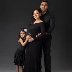 A classic family portrait as they await a new member. The family, dressed in coordinated black outfits, stands together with the older daughter hugging her mom's baby bump, excited to become a big sister.