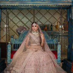 A regal shot of a model seated on a traditional blue swing, wearing a magnificent pink bridal lehenga with a sheer veil.