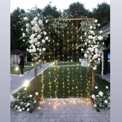 A simple and elegant photo booth perfect for an evening reception. The wooden frame is adorned with white roses and greenery, with a curtain of warm fairy lights creating a magical backdrop.