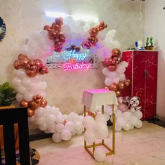 A classy rose gold and white balloon ring backdrop. This setup, complete with a neon sign and a pedestal for the cake, is perfect for an elegant birthday celebration at home.