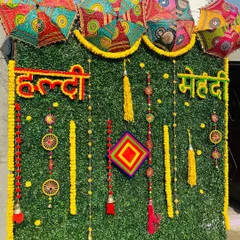 For a modern take on traditional Haldi and Mehndi decor, I use an artificial green wall as a base. This backdrop is decorated with marigold garlands, colorful tassels, and Rajasthani umbrellas to create a vibrant and festive photo corner.