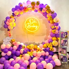 A vibrant baby shower decoration with a purple, pink, and gold balloon arch. The setup includes an 'Oh Baby' neon sign and transparent 'BABY' blocks, creating a beautiful photo corner.