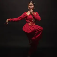 A beautifully frozen pose from a classical dance session. The studio lighting is carefully positioned to sculpt the dancer's form and highlight the rich red of her traditional costume against the dark background.