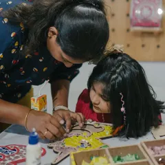 A mother helps her daughter with a mosaic project, sharing a quiet moment of focus and collaboration. Our studio is a space for creating these beautiful, shared experiences.