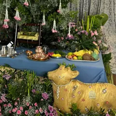 A wider view of the Janmashtami setup, showing the golden cow prop in context. The altar is adorned with fresh fruit, copper utensils, and a miniature swing, all surrounded by abundant flowers and greenery.