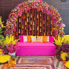 A stunning Mehendi seating area with a dense floral arch of pink and yellow flowers. The backdrop features hanging tassels, and the space is decorated with colorful cushions and a woven rug.