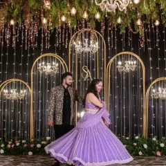 A couple enjoying their Sangeet night on a beautifully lit stage. The backdrop of fairy lights and golden arches, combined with hanging chandeliers, creates a magical and romantic setting for their performance.