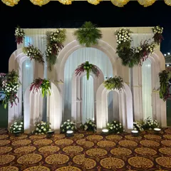 A beautiful stage backdrop with layered white arches and abundant white and burgundy floral arrangements, accented by spotlights for a dramatic evening effect.