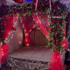 A closer look at the bed decoration, featuring a lush arch of fresh red roses and green leaves over the headboard. The arch is intertwined with delicate fairy lights and draped with red tulle for a soft, romantic feel.
