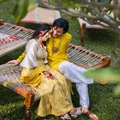 A quiet, romantic moment during their Haldi ceremony. The couple, dressed in yellow, share a tender moment on a traditional charpai, surrounded by lush greenery.