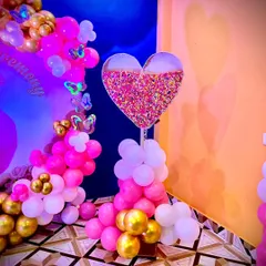 A unique decorative piece for a naming ceremony. This heart-shaped stand is filled with colorful sprinkles and surrounded by pink, white, and gold balloons.