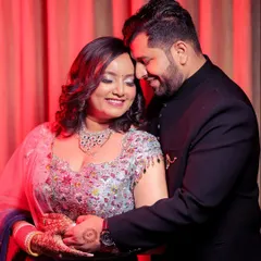 This is a warm, romantic portrait from an engagement celebration. The couple holds each other in a gentle embrace, their smiles showing their comfort and love. The red curtain backdrop adds a touch of drama and richness to the scene.