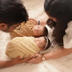 An intimate, top-down view of parents admiring their sleeping newborn twins. The babies are swaddled in complementary colors, creating a warm and harmonious family portrait.