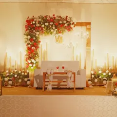 A romantic and warm stage setting perfect for an anniversary or reception. The design uses an L-shaped floral arrangement of red and white roses, complemented by numerous candles for soft lighting.