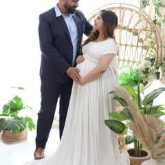 This portrait showcases a light and airy studio setup with natural elements like pampas grass and plants. The couple shares a loving gaze, with the mother-to-be glowing in a flowing white empire-waist gown.