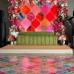 This vibrant Mehendi stage features a colorful Moroccan tile backdrop, framed by a lush arch of pink and orange flowers, and a modern olive green sofa.