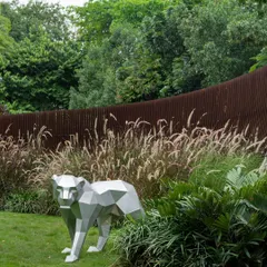 A geometric, silver-colored bear sculpture stands in a grassy area, with a curving Corten steel screen in the background. This juxtaposition of art and landscape creates a playful yet sophisticated focal point.