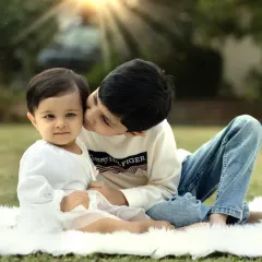 A sweet moment between siblings outdoors, where an older brother gives his baby sister a gentle kiss on the cheek under the warm sun.