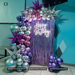 A vibrant setup with a purple fringe backdrop and a neon "Happy Birthday" sign. The balloon arch in shades of purple and silver, with starburst foil balloons, creates a futuristic, high-energy vibe.