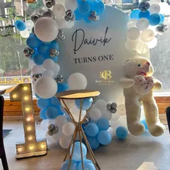 A sweet and simple setup for a first birthday. The 'Daivik Turns One' theme features a personalized backdrop, a blue and white balloon garland, a light-up number '1', and a cute teddy bear prop.