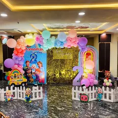 A complete Cocomelon birthday setup in a party hall. This decoration features a large backdrop with all the Cocomelon characters, a gold sequin panel, character cutouts, and a pastel rainbow balloon arch.