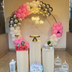 Here is a fun and vibrant bachelorette party setup. The centerpiece is a circular frame with a multi-colored balloon arch in pink, white, and silver chrome. A 'Bride to Be' neon sign adds a fun glow, while decorative plinths, lanterns, and birdcages complete the theme.