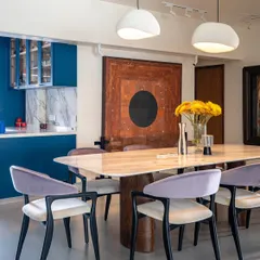 The dining area is designed for memorable gatherings, featuring a striking marble-topped table with sculptural wooden legs. We used lavender-hued chairs to introduce a soft, sophisticated color that complements the bold blue of the adjacent open kitchen.