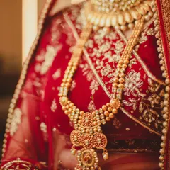 This shot focuses entirely on the magnificent gold temple jewelry worn by the bride. The warm lighting enhances the glow of the gold, highlighting the intricate patterns of her necklace against the red silk of her lehenga.