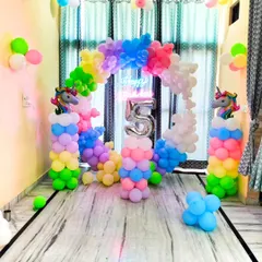 A full view of the unicorn-themed birthday decoration at home. The setup includes a circular balloon arch in pastel rainbow colors, a neon sign, and a number "5" foil balloon, transforming the living room into a fantasy land.