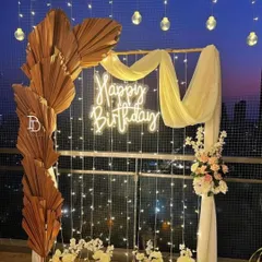 An elegant balcony decoration with a 'Happy Birthday' neon sign as the centerpiece. The setup is framed by dried palm leaves, draped fabric, fairy lights, and flameless candles, creating a magical evening ambiance.