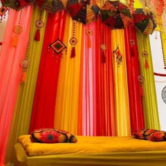 A close-up of a colorful Mehandi seating area. The decor features a low gadda with printed cushions and a backdrop of bright drapes with hanging Rajasthani umbrellas.