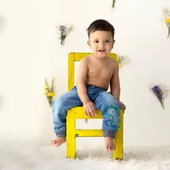 A happy baby boy in jeans sits confidently on a bright yellow chair. The simple floral background adds a touch of whimsy to this cheerful sitter portrait, capturing his bright and happy spirit.