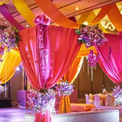 We use a combination of vibrant drapery and floral arrangements to create a festive sangeet or reception stage. The colorful ceiling drapes in pink, orange, and yellow complement the purple orchid bouquets on each pillar.
