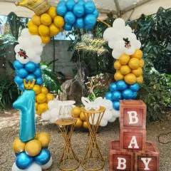 A royal-themed first birthday for a little prince. This outdoor setup includes a blue, white, and gold balloon arch topped with a crown, a large number '1' balloon, 'BABY' blocks, and a light-up name sign.