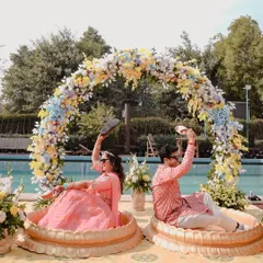 A fun and quirky shot from a poolside Haldi ceremony. The couple playfully holds up slippers, showing their fun-loving personalities against a beautiful floral arch.