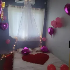 A simple and romantic first-night bedroom decoration, featuring a canopy with fairy lights, heart-shaped balloons, and a heart made of rose petals on the bed.