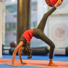 A student executes a graceful bridge pose, lifting one leg to touch a stability ball. This drill improves back flexibility, core strength, and body awareness, key elements in artistic gymnastics.