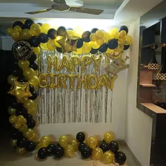 A black and gold birthday decoration for a home party. This setup includes a balloon arch, 'Happy Birthday' foil letters, and star-shaped foil balloons.