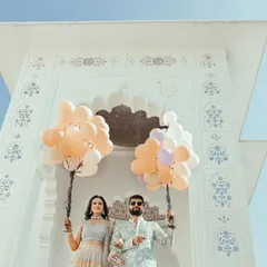 A creative low-angle shot of the couple holding balloons, framed by a beautiful white archway, capturing the fun carnival theme of their pre-wedding event.